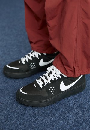 Baskets Nike noir et blanc avec lacets blancs portées avec un pantalon rouge ample, debout sur un sol recouvert de moquette bleue.