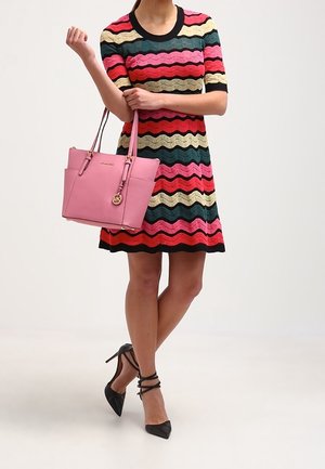 Femme en robe rayée multicolore ondulée tenant un sac à main rose, portant des talons hauts noirs à brides, debout les jambes croisées.