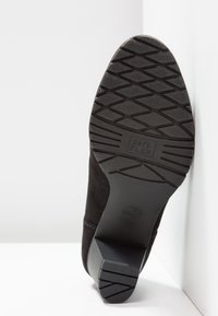 Semelle de chaussure noire avec motif antidérapant texturé et talon bloc, étiquetée soft flex, inclinée contre un mur et un sol blancs.