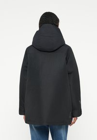 Άτομο με μαύρο φούτερ με κουκούλα και μακριά μανίκια, φορόντας μπλε τζιν, στραμμένο μακριά σε έναν απλό λευκό φόντο.