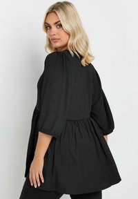 Top nero con maniche a sbuffo e vita arricciata. Tessuto testurizzato, vestibilità rilassata, design di media lunghezza. La vista posteriore mostra la pienezza dell'orlo.