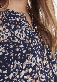 Marinblus med ett blommönster i vitt och ljusbrunt. Har en knapplist och liten detalj vid halsringningen. Mjuk, texturerad tyg.