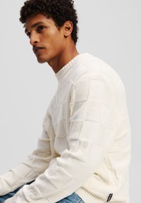 Weißer Strickpullover mit einem strukturierten, gitterartigen Muster. Verfügt über einen gerippten Rundhalsausschnitt und wird mit blauen Denim-Jeans kombiniert.