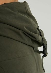 Primer plano de una chaqueta con capucha verde que muestra la textura de la tela y una hebilla de plástico negro con cordones ajustables.