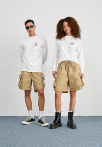 Hvit langermet skjorte med svart trekantet logo, beige cargo shorts og kontrastfargede svarte militærstøvler. Tilbehør inkluderer solbriller og kjede.