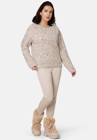 Strickpullover in Creme mit bunten Sprenkeln, entspannte Passform, lange Ärmel, kombiniert mit beigen Leggings und beigen, fellgefütterten Stiefeln.
