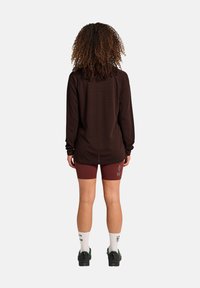 Braunes Langarmshirt, kastanienbraune Shorts mit ‚ATW‘-Logo, weiße Socken mit Text und schwarze Sportschuhe. Lockige Haare sind von hinten sichtbar.