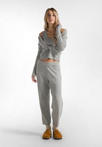Grijze gebreide cropped trui met een gedraaide voorkant en lange mouwen, gecombineerd met bijpassende grijze sweatpants en bruine instappers.