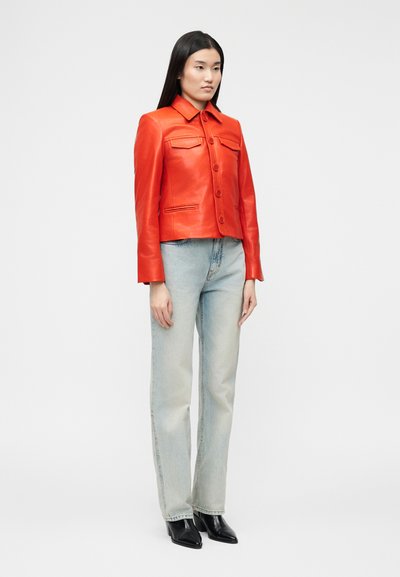 Γυναίκα στέκεται φορώντας ένα φωτεινό κόκκινο δερμάτινο μπουφάν, ανοιχτό μπλε τζιν και μαύρα μποτάκια με τακούνι μπροστά σε ένα απλό φωτεινό φόντο.