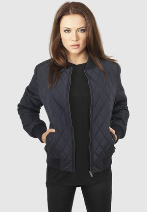 Femme aux cheveux bruns raides portant une veste bomber matelassée bleu marine sur des vêtements noirs, mains dans les poches de la veste, fond neutre.