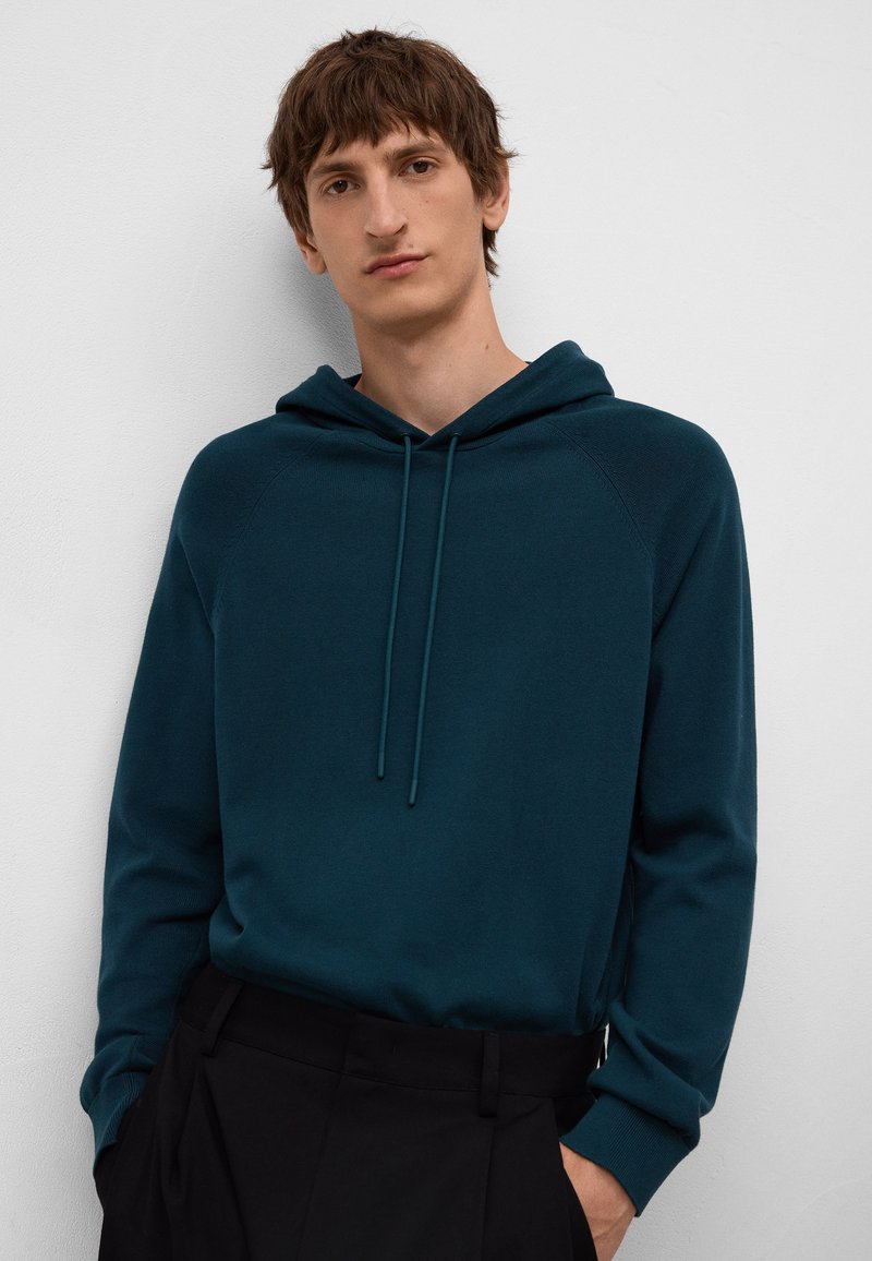 Felpa teal con cappuccio e cordini, maniche raglan e polsini a costine, abbinata a pantaloni neri. Tessuto morbido con una vestibilità rilassata.