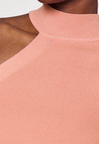 Top de punto acanalado en color durazno con un alto cuello y un diseño asimétrico en el hombro, que presenta una textura suave y un detalle sutil.