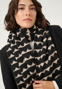 Sciarpa nera e beige con un motivo geometrico, realizzata in un morbido materiale testurizzato, appoggiata attorno al collo, coprendo parzialmente una giacca nera.