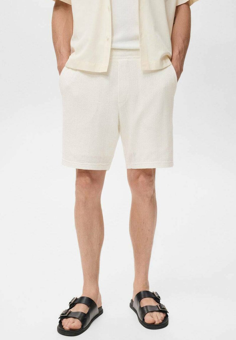 Shorts blancs texturés avec une taille élastique, présentant des poches latérales. Associés à des sandales noires avec deux brides à boucle.
