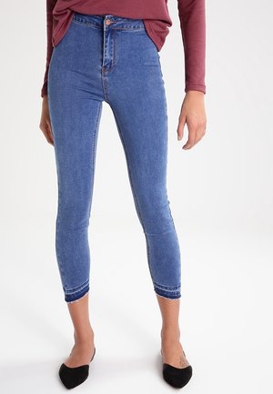 Donna che indossa jeans skinny blu a vita alta e corti con orli sfrangiati, abbinati a ballerine nere con punta e una maglia a maniche lunghe color marsala.