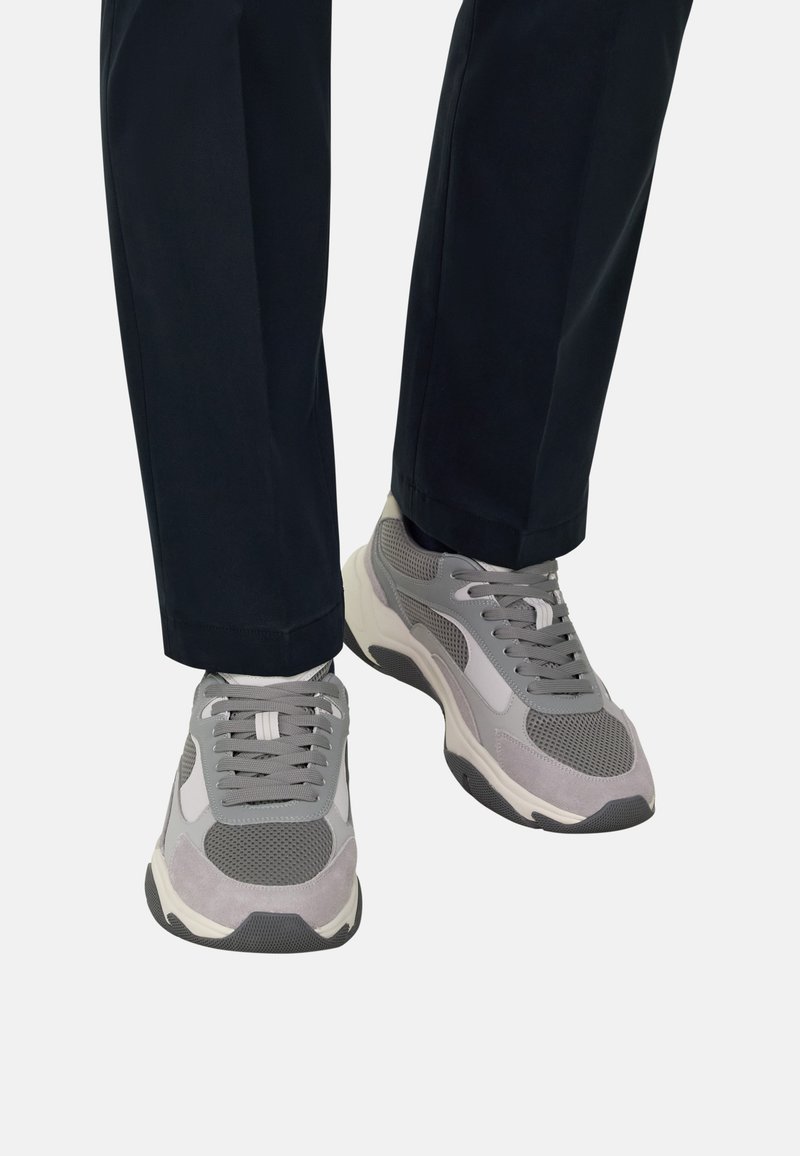 Paire de baskets grises et blanches portées avec un pantalon noir, posées sur un fond blanc uni.