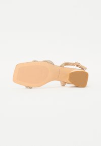 Sandale nude à talons avec une sangle arrière texturée et des accents décoratifs en strass. La semelle en caoutchouc présente des lignes de grip horizontales.