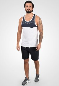 Débardeur blanc avec une section supérieure à motif marine et une poche, associé à des shorts noirs et des baskets grises avec des accents blancs.