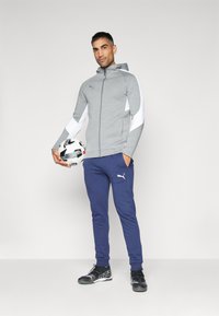 Grå sportjakke med hvide detaljer, parret med blå bukser. Personen holder en fodbold. Sorte træningssko fuldender outfittet.