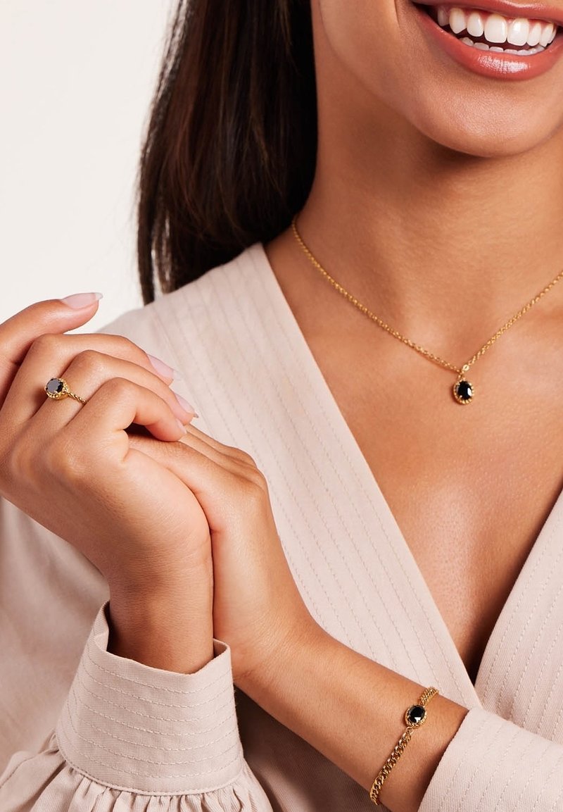 Gouden sieraden omvatten een ketting en armband met zwarte edelstenen. De ring heeft een vergelijkbaar gevormde zwarte steen. Het model draagt een lichte, gestructureerde blouse.