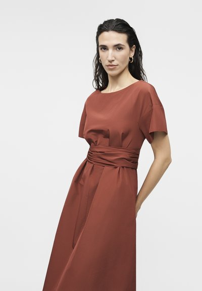 Γυναίκα με φορέμα κοντό μανίκι σε σκουριά χρώμα, με φαρδιά, σούρες στη μέση και χρυσούς κρίκους στα αυτιά, στέκεται μπροστά σε ανοιχτό φόντο.