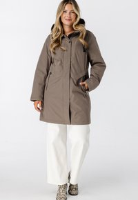 Bruine, geïsoleerde lange jas met een capuchon, rits aan de voorkant en zijzakken; gecombineerd met witte wijde broek en laarzen met slangenprint.