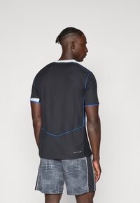 Maglietta sportiva nera con tessuto testurizzato, presenta dettagli blu lungo le cuciture e un colletto bianco. Abbinata a pantaloni corti grigi a fantasia.