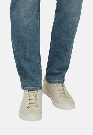 Person trägt hellbeige Leder-Sneaker mit Schnürsenkeln und blaue Jeans aus Denim, steht vor einem schlichten weißen Hintergrund.