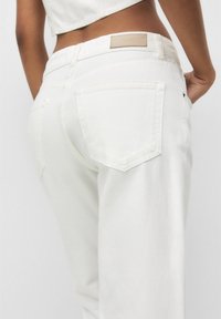 Jean en denim blanc à coupe décontractée, avec deux poches arrière et une patch en cuir beige marqué sur la taille. Texture lisse.