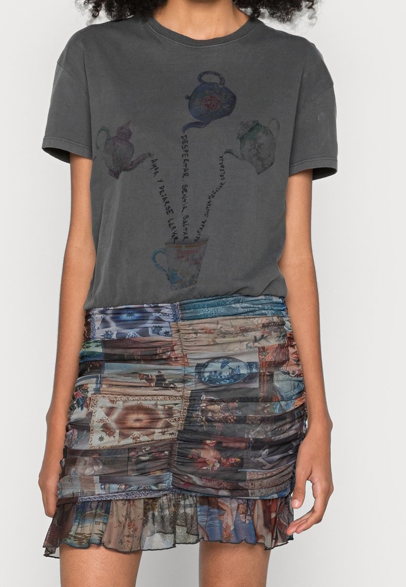Jeune personne portant un t-shirt gris foncé avec des illustrations de théière et de tasse ainsi qu'une jupe froncée multicolore avec des motifs imprimés.