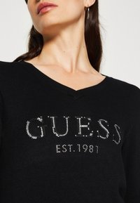 Zwarte v-hals trui met een met rhinestones versierde "GUESS" logo en "EST. 1981" detail, met een zachte, textuurlichte stofafwerking.