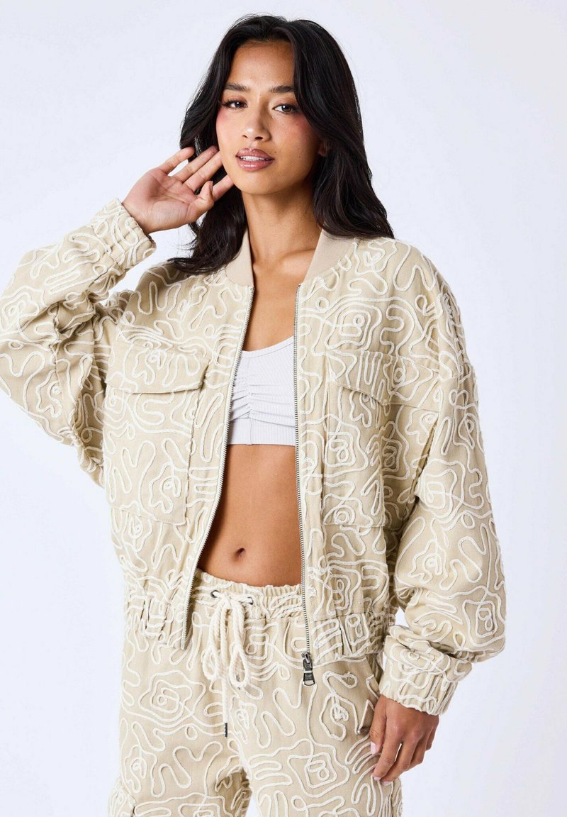 Veste bomber en khaki avec broderie abstraite blanche, col et poignets côtelés, dotée de poches avant et d'une fermeture éclair.