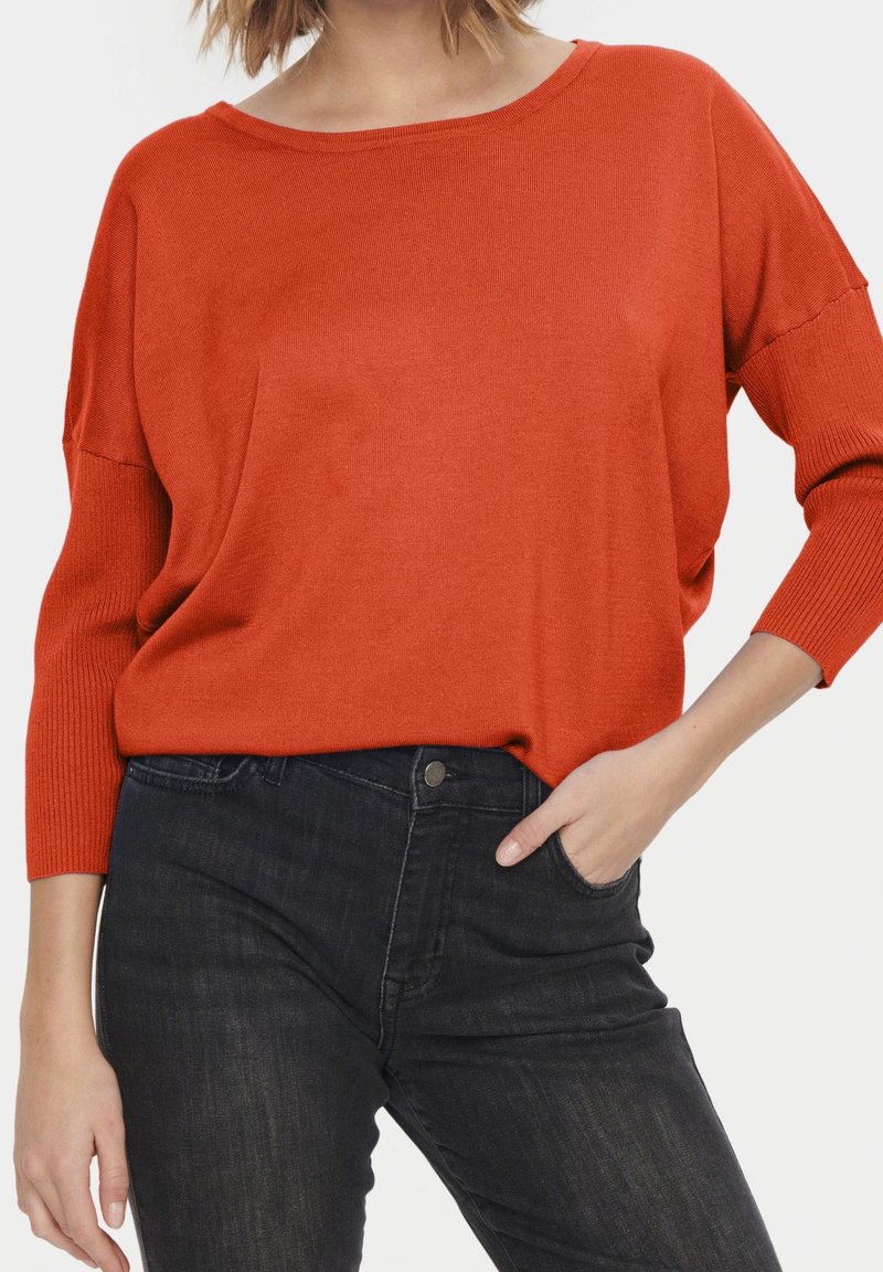 Femme portant un pull orange ample à manches longues avec un jean délavé foncé, une main reposant dans la poche, sur fond blanc.
