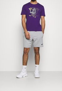 Fialové tričko s krátkým rukávem a grafickým potiskem, spárované se šedými šortkami. Bílá sneakers a ponožky s logem dokončují vzhled.
