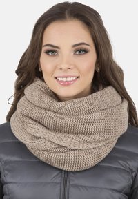 Sciarpa infinita lavorata a maglia di un beige chiaro, con un motivo spesso e testurizzato. Indossata sopra un giubbotto imbottito scuro, offre una calda comodità.