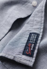 Grijs-blauwe katoenen shirtstof met fijne textuur, opstaande kraag en een zichtbare label met rode en marineblauwe tekst.