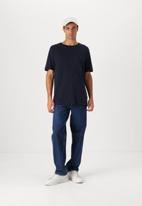 Marineblå t-skjorte med kort erm og lomme, kombinert med løstsittende blå jeans og hvite sneakers. Modellen har på seg en lyseferdig caps.