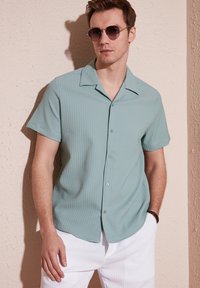 Lichtgroene geribde shirt met korte mouwen en een notch-kraag, knoopsluiting aan de voorkant en subtiele textuur; gedragen met witte broek en zonnebril.
