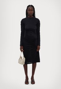 Zwarte lange mouwen jurk met gerimpelde details, tot halverwege de kuit. Het model houdt een metalen mesh handtas vast en draagt strappy zwarte sandalen.