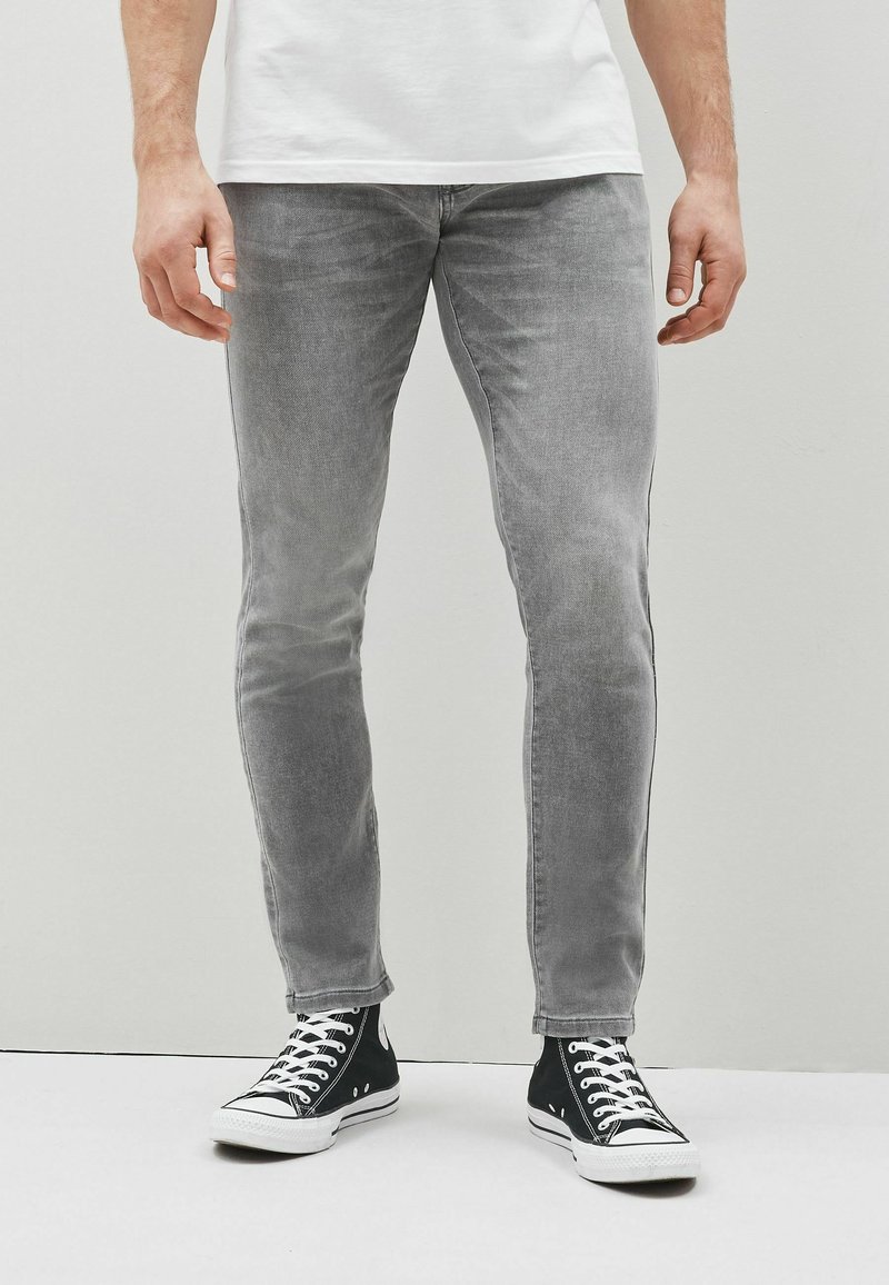 Bărbat îmbrăcat în blugi slim-fit gri deschis, adidași negri cu șireturi albe și un tricou alb, stând în fața unui perete simplu de culoare deschisă.