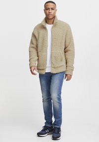 Beige fleecejakke med høy krage, frontlommer og glidelås. Kombinert med blå jeans og marineblå sneakers. Solide teksturer gjennomgående.