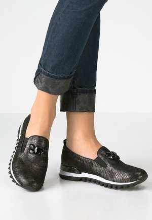 Osoba nosząca czarne teksturowane buty wsuwane z ozdobnymi czarnymi dodatkami oraz ciemne podwinięte dżinsy na jednolitym jasnym tle.