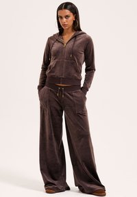 Bruine velours trainingspak met ritsjack met capuchon en wijde broekspijpen. Voorzien van goudkleurige hardware, zijzakken en contrasterende tailleband.