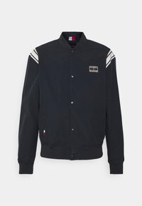 Giacca bomber nera con chiusura a pressione, polsini a costine e due strisce bianche sulle spalle. Presenta una patch con logo sul petto.