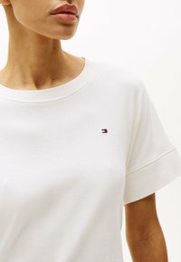 T-shirt en coton blanc avec un col rond et des manches courtes. Présente un petit logo brodé en rouge, blanc et bleu sur la poitrine.