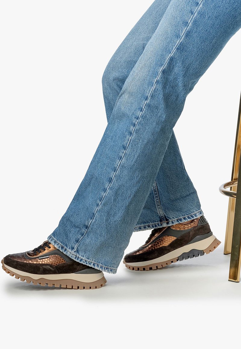 Personne portant un jean bleu et des baskets texturées marron avec de épaisses semelles beiges, assise sur un tabouret métallique doré.