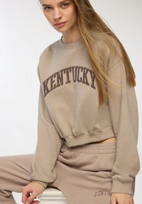 Felpa corta beige con "KENTUCKY" stampato in marrone scuro, abbinata a pantaloni della tuta beige coordinati, con vita elastica.
