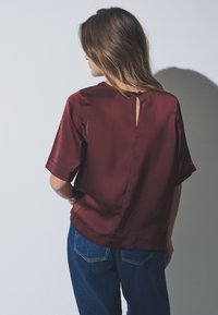 Bordeauxrote Satinbluse mit kurzen Ärmeln, lockerem Schnitt und einer Schlitzöffnung am Rücken. Kombiniert mit blauen Jeans aus Denim.