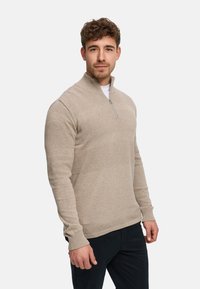 Beige stickad pullover med halvlång dragkedjestor krage, med texturerade mönster över kroppen och ribbstickade ärmslut och fåll.