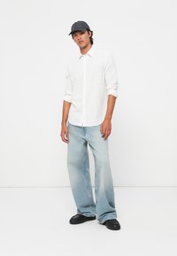 White long-sleeve button-up shirt, light blue wide-leg jeans, and black shoes, complemented by a dark cap. Simple, clean design.